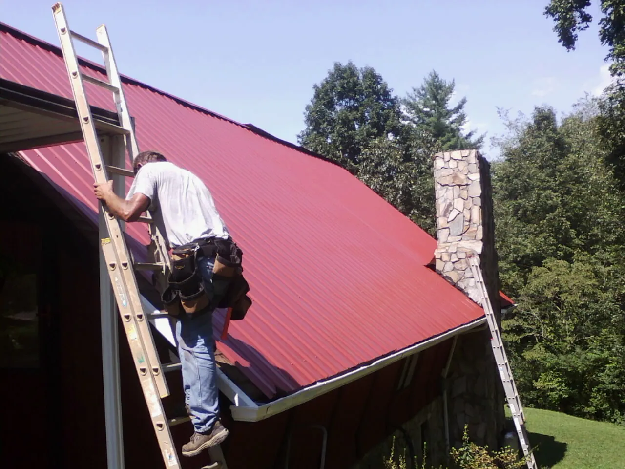 Roofer on a ladder beside a red metal roof and stone chimney — Soffit & Fascia Repair by Roofing Brothers in Valley View